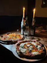 A moody, low-light shot of two pizzas served on metal trays at a table illuminated by two candles in wine bottles. The setting is intimate and atmospheric, typical of a pub or casual dining venue.