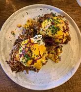 A top-down, overhead shot of a pulled pork benedict brunch dish served on a rustic plate, featuring poached eggs, hollandaise sauce, and a side of sweetcorn salsa.