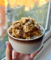 A close-up, eye-level shot of a bowl of tofu and leeks served over rice, held by a hand in front of a window. The dish is garnished with sesame seeds and appears to be a home-cooked meal.