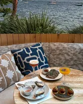 A high-quality, well-composed shot of a table spread at a waterside venue, featuring oysters, cocktails, and small plates. The natural lighting and scenic background create an inviting atmosphere that effectively showcases the venue's aesthetic and menu.