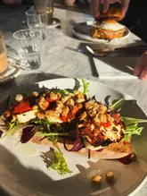 A close-up, top-down shot of a halloumi sandwich on a plate, with a burger visible in the background. The scene is captured in direct, harsh sunlight, creating high-contrast shadows on the food and table.