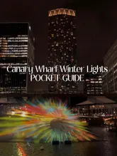 A visually striking night-time shot of a light installation at Canary Wharf, serving as a cover image for a guide to the Winter Lights event. The composition effectively frames the colourful projection against the dark, illuminated city skyline.