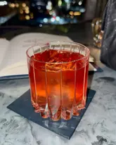 A high-quality, moody shot of a cocktail in a premium bar setting. The image features a well-lit cocktail with a large ice cube, set against a blurred, atmospheric background that suggests an upscale venue.