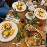 A top-down flat lay photograph of a brunch spread featuring plates of eggs benedict served on croissant swirls, a skillet dish with toasted bread, and hot drinks, with red roses arranged diagonally across the table.