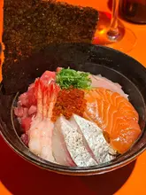 A close-up, overhead shot of a chirashi sushi bowl featuring various types of sashimi, shrimp, and roe, served in a dark bowl with a sheet of nori.