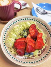 A bright, overhead shot of a fresh acai bowl topped with sliced banana, strawberries, blueberries, and a drizzle of sauce, accompanied by a latte.