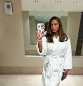 A mirror selfie taken in a hotel or spa bathroom, featuring a woman wearing a white bathrobe.
