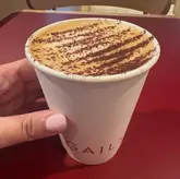 A close-up, eye-level shot of a takeaway coffee cup from Gail's Bakery held by a hand against a plain red table surface.