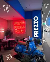 A wide-angle shot of a Prezzo restaurant interior, featuring a blue Vespa scooter as a decorative prop and a prominent red neon sign reading 'PASTA PIZZA VINO'. The image is taken from the perspective of a shopping mall corridor, looking into the dining area.