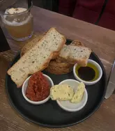 A simple, overhead shot of a bread board starter featuring rustic bread, wild garlic butter, tomato tapenade, and balsamic oil, accompanied by a cocktail in the background.