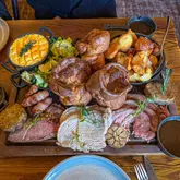 A high-quality, top-down shot of a substantial Sunday roast sharing board, featuring a trio of meats including sirloin beef, leg of lamb, and turkey, accompanied by Yorkshire puddings, roast potatoes, pigs in blankets, stuffing, and sides served in cast iron dishes. The lighting is natural and even, effectively highlighting the textures of the food.