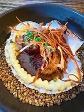 A close-up, top-down shot of a scallop dish served in a shell, featuring parsnip puree and crisps, presented on a bed of grains.