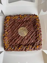 A high-angle, well-lit shot of a square brownie slab presented in a white takeaway box. The brownie is decorated with a neat chocolate drizzle and gold and silver sprinkles, featuring a central 'Ramadan Kareem' topper.