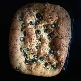 A top-down, overhead shot of a freshly baked focaccia bread topped with black olives and feta cheese, presented against a dark, plain background.