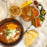 A high-quality, overhead flat-lay photograph of a brunch spread featuring a shakshuka, a Turkish breakfast platter, and an iced latte on a marble table.