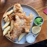 A top-down, casual shot of a classic fish and chips meal served on a wooden table. The image captures the texture of the battered fish and the accompanying sides, including minted peas and tartar sauce, typical of a British pub setting.