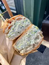 A close-up, handheld shot of a chicken sandwich served in a paper bag, likely taken at a cafe or street food venue. The focus is sharp on the texture of the filling and the bread, highlighting the freshness of the ingredients.