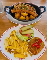 A top-down shot of two dishes from a restaurant, featuring a sausage plate with roast potatoes and a halloumi schnitzel burger with fries.