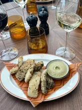 A top-down shot of a plate of fried food, likely halloumi fries, served with a dip on a restaurant table. The table is set with multiple wine glasses and condiments.