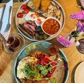 A high-quality, overhead flat-lay shot of a brunch spread featuring a Full English breakfast and a chicken waffle, accompanied by tea and hot chocolate. The lighting is bright and natural, highlighting the textures of the food, and the composition is clean and well-balanced.