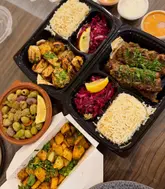 A top-down, flat-lay photograph of a Lebanese takeaway spread, featuring containers of lamb kafta, shish taouk, rice, salad, olives, and batata harra on a wooden table.