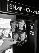 A black and white, first-person perspective shot showing a hand holding a photo strip from a photo booth at Spinners. The image highlights the 'Snap-O-Matic' photo booth experience, focusing on the physical souvenir from a night out.