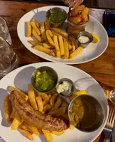 A top-down, overhead shot of two plates of pub-style food served on a wooden table. One plate features a large sausage with chips and a side of curry sauce, while the other contains scampi, chips, and mushy peas.