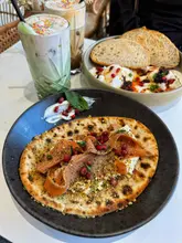 A vibrant, well-composed overhead shot of a brunch spread featuring a roasted carrot and dukkah flatbread, a bowl of Cilbir, and two iced drinks. The lighting is soft and natural, highlighting the textures and colours of the food.