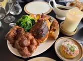 A high-quality, appetizing close-up shot of a Sunday roast dinner featuring a whole roast chicken, large Yorkshire puddings, and greens, accompanied by a side of potatoes and a gravy boat, set on a table with a lit candle.