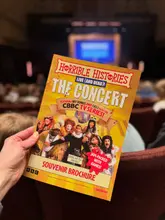 A first-person perspective shot holding a 'Horrible Histories: The Concert' souvenir brochure inside a theatre, likely the Theatre Royal Nottingham. The image serves as a visual anchor for a review of the live show, highlighting the family-friendly nature of the event.