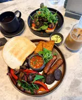 A high-quality, overhead flat-lay photograph of a brunch spread at Fearns, featuring a vegan breakfast plate with sausages, black pudding, hash browns, beans, and sourdough, alongside a mushroom sourdough dish, an iced latte, and an Americano.