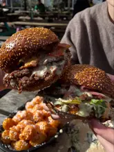 A close-up, handheld shot of two loaded smash burgers being held up in front of a pub beer garden setting, with a side of loaded fries visible in the foreground.