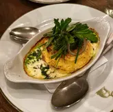 A close-up, eye-level shot of a cheese soufflé served in an oval dish, garnished with fresh herbs and red onion, presented on a white plate with a spoon.