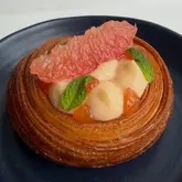 A high-quality, close-up food photograph of a grapefruit and mint croissant tart served on a dark plate. The image highlights the intricate lamination of the pastry and the vibrant colors of the curd and fruit topping.