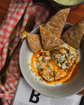 A high-quality, top-down photograph of Turkish eggs served with toasted pitta bread, presented on a rustic surface with a red checkered napkin and a book underneath.