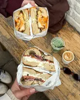 A high-quality, lifestyle-focused shot of two loaded bagels being held over a wooden table, accompanied by a matcha latte and a coffee. The image captures a casual, aesthetic brunch setting, likely at a bakery or cafe.