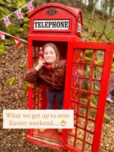 A candid, family-oriented lifestyle shot featuring a young girl posing inside a red telephone box located in a wooded area, likely part of an Easter-themed day out. The image captures a relaxed, seasonal moment with festive bunting visible in the background.