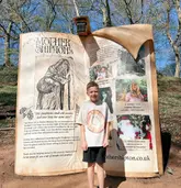 A photo of a young boy standing in front of a large, book-shaped information board at Mother Shipton's Cave, highlighting the venue's themed events.