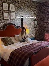 A promotional image for a hotel stay in North Berwick, featuring a creator in a bathrobe on a bed with a glass of champagne, set against a highly patterned wallpaper background.