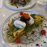 A high-quality, overhead shot of a brunch dish featuring avocado toast topped with poached eggs, halloumi, microgreens, and edible flowers, served on a clean, neutral table surface.