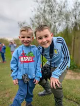 A high-quality, candid photograph of two young boys smiling at the camera while outdoors, wearing binoculars, suggesting a nature-based activity or exploration experience.