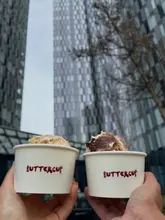 A handheld shot of two cups of banana pudding held up against a backdrop of city skyscrapers in Manchester. The image captures the product in a casual, on-the-go setting.