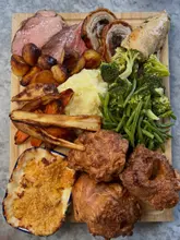 A top-down, overhead shot of a generous Sunday roast sharing board served on a wooden platter, featuring slices of beef, porchetta, chicken, roast potatoes, carrots, parsnips, green beans, broccoli, cauliflower cheese, and large Yorkshire puddings.