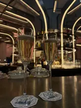 A high-quality, atmospheric shot of two glasses of champagne sitting on a bar counter, with the blurred, warm-lit interior of a bar in the background. The image captures a sophisticated, relaxing vibe, likely at a cocktail bar or lounge.