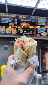 Here are a few options for your SEO alt text, all under 125 characters:

*   **Option 1 (Descriptive):** A delicious authentic Kurdish style shawarma wrap held in hand at Al Aqsa takeaway on Evington Road, Leicester.
*   **Option 2 (Action-focused):** Someone holding a fresh Al Aqsa Kurdish shawarma wrap from the local takeaway shop on Evington Road, Leicester.
*   **Option 3 (Concise):** Fresh Kurdish shawarma wrap from Al Aqsa, a popular halal takeaway spot on Evington Road in Leicester.