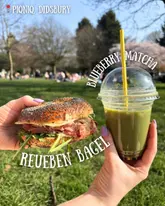 A bright, outdoor shot of a Reuben bagel and a blueberry matcha iced latte held in front of a blurred park background, effectively capturing a casual, sunny picnic vibe.