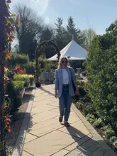 A bright, sunny lifestyle shot of a woman walking along a paved path in a garden centre, with plants and a marquee visible in the background.