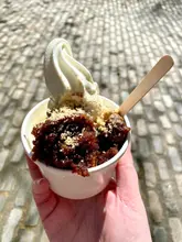 A close-up, handheld shot of a sticky toffee pudding topped with a swirl of soft serve ice cream, set against a cobblestone background.