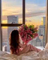 A high-quality, aspirational lifestyle shot featuring a person sitting on a bed in a hotel room, holding a large bouquet of pink tulips against a backdrop of a city skyline at sunset. The image effectively communicates a romantic or celebratory atmosphere.
