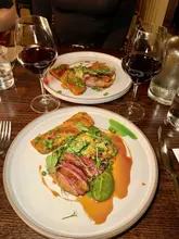 A top-down shot of a two-course meal featuring roasted duck breast with a pistachio rosti, accompanied by glasses of red wine. The setting appears to be a restaurant dinner service.